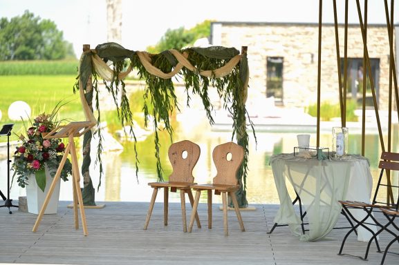 Freie Trauung am Wasser mit dekoriertem Traubogen aus Grün und Stoff, zwei Holzstühlen und Rednerpult.